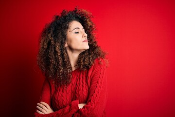 Young beautiful woman with curly hair and piercing wearing casual red sweater looking to the side with arms crossed convinced and confident