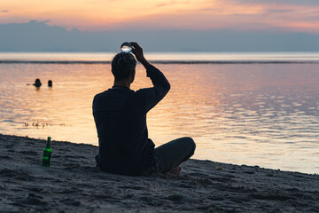 A man sits and tries to maintain balance with a transparent ball on his head on the beach at sunset