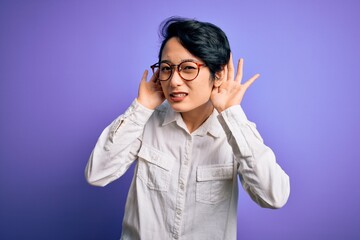 Young beautiful asian girl wearing casual shirt and glasses standing over purple background Trying to hear both hands on ear gesture, curious for gossip. Hearing problem, deaf