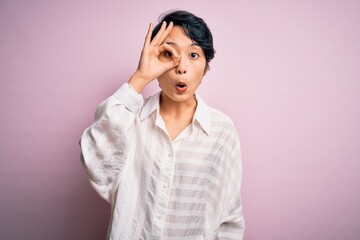 Young beautiful asian girl wearing casual shirt standing over isolated pink background doing ok gesture shocked with surprised face, eye looking through fingers. Unbelieving expression.