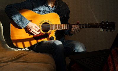 Man with acoustic guitar in a shirt and cap plays live staying home for the audience in front of his laptop  during quarantine and isolation lockdown due to coronavirus. Work from home concept.