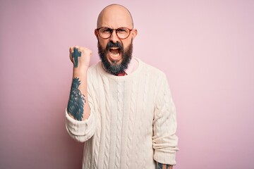 Handsome bald man with beard and tattoo wearing glasses and sweater over pink background angry and mad raising fist frustrated and furious while shouting with anger. Rage and aggressive concept.
