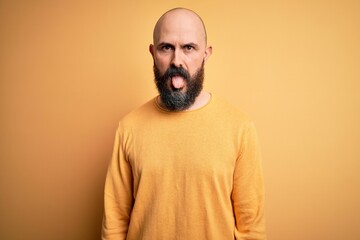 Obraz premium Handsome bald man with beard wearing casual sweater standing over yellow background sticking tongue out happy with funny expression. Emotion concept.