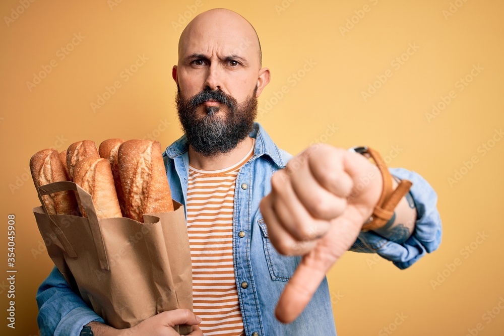 Poster Handsome bald man with beard holding paper bag with bread over yellow background with angry face, negative sign showing dislike with thumbs down, rejection concept