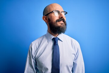 Handsome business bald man with beard wearing elegant tie and glasses over blue background looking away to side with smile on face, natural expression. Laughing confident.