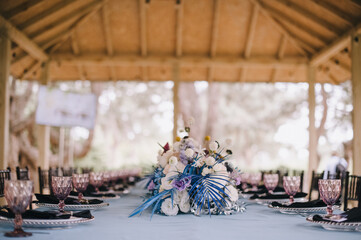 Naklejka premium Outdoor banquet table under the wooden roof of the gazebo. Rack and cutlery, purple glasses. Floral arrangement of violet, blue, black, silver flowers. Black banquet chairs. On the table is a blue tab