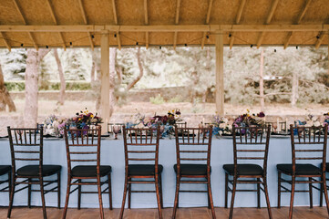 Outdoor banquet table under the wooden roof of the gazebo. Rack and cutlery, purple glasses. Floral arrangement of violet, blue, black, silver flowers. Black banquet chairs. On the table is a blue tab