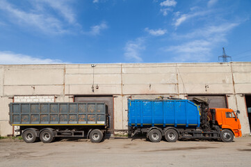 Large truck with orange cab, a blue body and a gray trailer against a concrete warehouse with an iron gate closed and the sky on a sunny day waiting for the cargo to be unloaded by a transport company