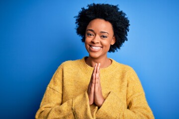 Young beautiful African American afro woman with curly hair wearing yellow casual sweater praying...