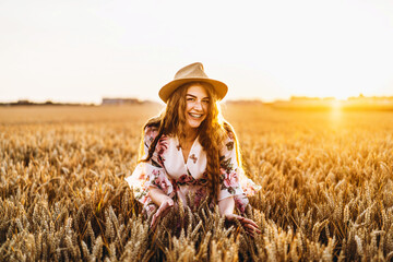 Young girl with long curly hairand freckles face, in hat, in light white dress with floral print, standing in wheat field, posing for camera, in background sundown.