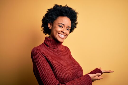 Young beautiful African American afro woman with curly hair wearing casual turtleneck sweater Inviting to enter smiling natural with open hand