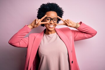 Young beautiful African American afro businesswoman with curly hair wearing pink jacket Doing peace symbol with fingers over face, smiling cheerful showing victory