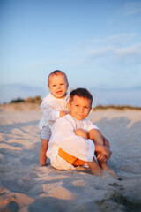 Two brothers in white linen shirts sit on the sand on the beach at sunset