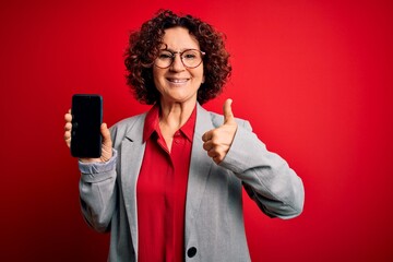 Middle age curly woman holding smartphone showing screen over isolated red background happy with big smile doing ok sign, thumb up with fingers, excellent sign