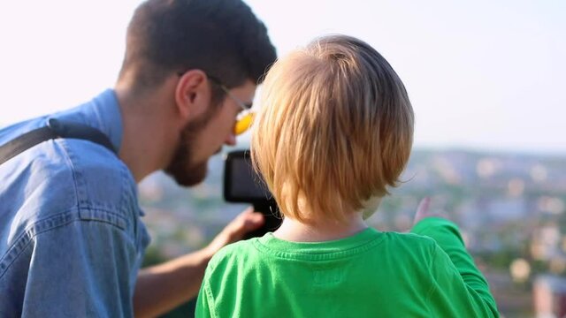Young father teaching little son to use modern professional camera, enjoying city landscape traveling together. Little boy learning using technology. Smart excited child exploring the horizon.