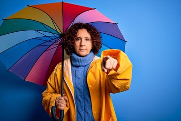 Middle age curly hair woman wearing rain coat holding colorful umbrella over blue background pointing with finger to the camera and to you, hand sign, positive and confident gesture from the front