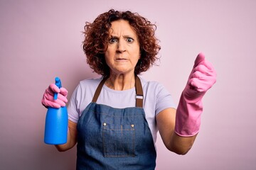 Middle age curly hair woman cleaning doing housework wearing apron and gloves using spayer annoyed and frustrated shouting with anger, crazy and yelling with raised hand, anger concept