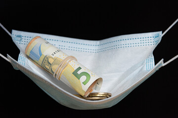 Medical face mask holding money inside of it, on a black background. World coronavirus economic losses concept. Concept of financial impact of covid-19 on European money area. 5 Euros and coins.