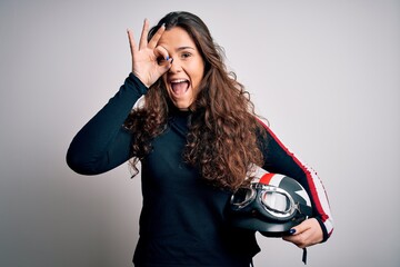 Beautiful motorcyclist woman with curly hair holding moto helmet over white background with happy face smiling doing ok sign with hand on eye looking through fingers