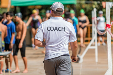 Back of male swimming coach walking on the poolside toward his young swimmers