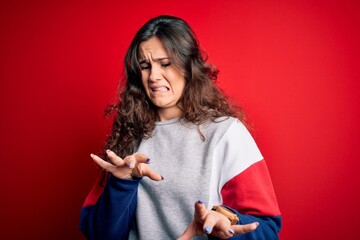 Young beautiful woman with curly hair wearing casual sweatshirt over isolated red background disgusted expression, displeased and fearful doing disgust face because aversion reaction.