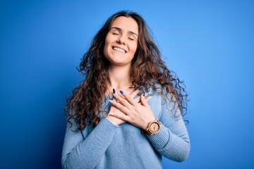 Young beautiful woman with curly hair wearing blue casual sweater over isolated background smiling with hands on chest with closed eyes and grateful gesture on face. Health concept.