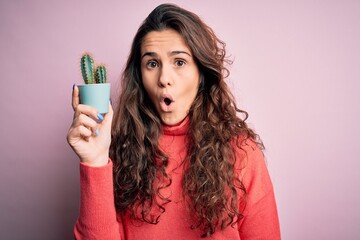 Young beautiful woman with curly hair holding small cactus over isolated pink background scared in shock with a surprise face, afraid and excited with fear expression