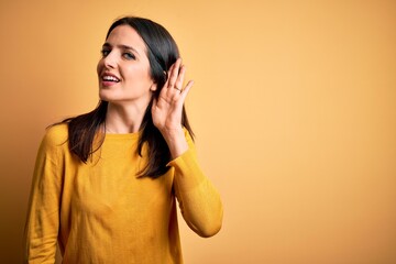Fototapeta premium Young brunette woman with blue eyes wearing casual sweater over yellow background smiling with hand over ear listening an hearing to rumor or gossip. Deafness concept.