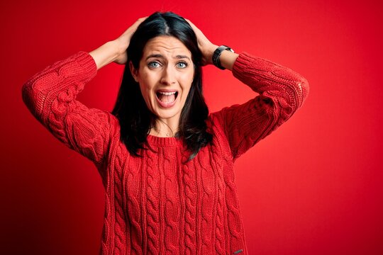 Young brunette woman with blue eyes wearing casual sweater over isolated red background Crazy and scared with hands on head, afraid and surprised of shock with open mouth