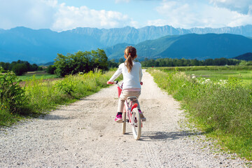 girl rides a pink bike on a dirt doron against a beautiful mountain landscape, view from the back - Powered by Adobe