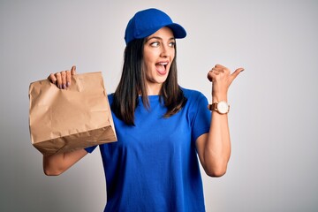 Young delivery woman with blue eyes wearing cap holding paper bag with food pointing and showing with thumb up to the side with happy face smiling