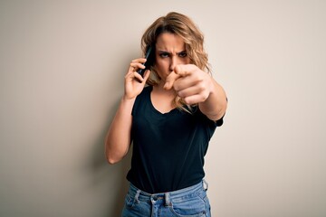 Beautiful blonde woman having conversation talking on the smartphone over white background pointing with finger to the camera and to you, hand sign, positive and confident gesture from the front