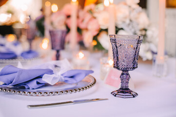 Presidium, banquet table with a white tablecloth. Plates and cutlery, purple glasses, napkins, candles in glass candlesticks. Bouquet of flowers, asters, roses, carnations, eustomas.