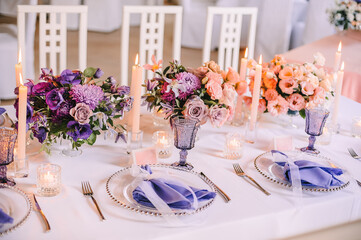 Presidium, banquet table with a white tablecloth. Plates and cutlery, purple glasses, napkins, candles in glass candlesticks. Bouquet of flowers, asters, roses, carnations, eustomas.
