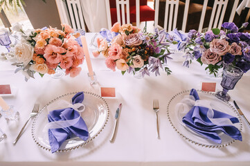 Presidium, banquet table with a white tablecloth. Plates and cutlery, purple glasses, napkins, candles in glass candlesticks. Bouquet of flowers, asters, roses, carnations, eustomas.