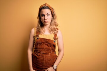 Young beautiful blonde woman wearing overalls and diadem standing over yellow background skeptic...