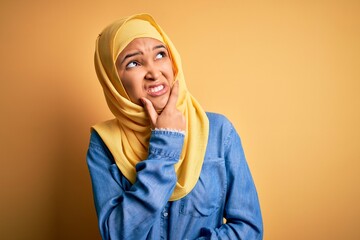 Fototapeta premium Young beautiful woman with curly hair wearing arab traditional hijab over yellow background Thinking worried about a question, concerned and nervous with hand on chin