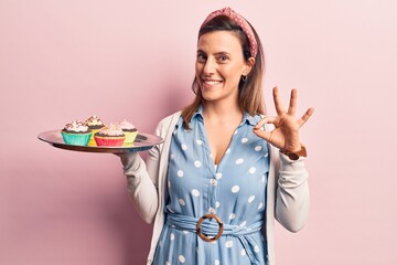 Young beautiful woman holding cupcake doing ok sign with fingers, smiling friendly gesturing excellent symbol