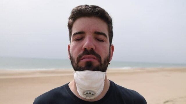 Young caucasian man with his eyes closed takes off his mask on the beach. Global pandemic due to coronavirus. New normality and life in open spaces and outdoors. Protection from COVID-19.