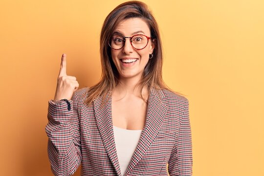 Young beautiful woman wearing business clothes and glasses pointing finger up with successful idea. exited and happy. number one.