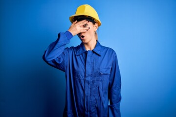 Young beautiful worker woman with blue eyes wearing security helmet and uniform peeking in shock covering face and eyes with hand, looking through fingers with embarrassed expression.