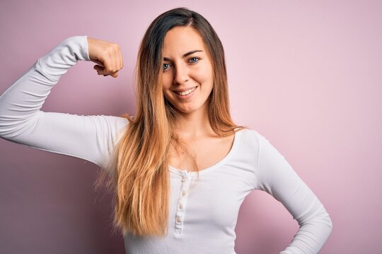 Young beautiful blonde woman with blue eyes wearing white t-shirt over pink background Strong person showing arm muscle, confident and proud of power