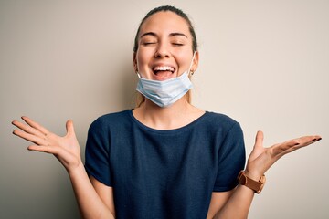 Beautiful blonde woman with blue eyes wearing medical mask over white background celebrating mad and crazy for success with arms raised and closed eyes screaming excited. Winner concept
