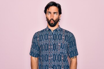 Young handsome hispanic bohemian man wearing hippie style over pink background making fish face with lips, crazy and comical gesture. Funny expression.