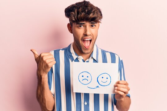 Young hispanic man holding sad to happy emotion paper pointing thumb up to the side smiling happy with open mouth
