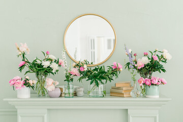 Pink and white peonies on the bookshelf  in glass vases. Round golden mirror on the wall above the fireplace