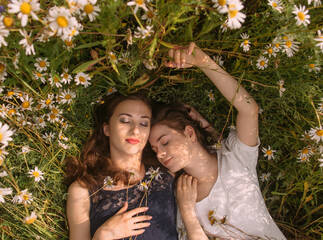 Two girls with closed eyes in dark blue and white dresses in sunny day lying down in chamomile field - 354058923