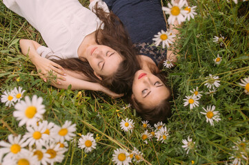Two girls with closed eyes in dark blue and white dresses in sunny day lying down  and sleeping in chamomile field - 354059139