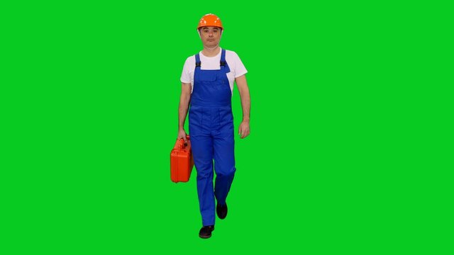 Confident asian builder in blue overalls and hard hat walking with red tools suitcase on green screen background, Chroma key 