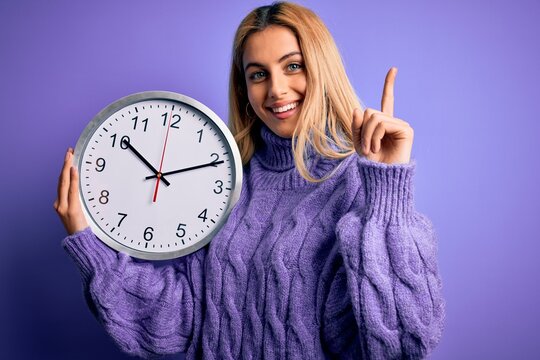 Young beautiful blonde woman doing countdown holding big clock over purple background surprised with an idea or question pointing finger with happy face, number one